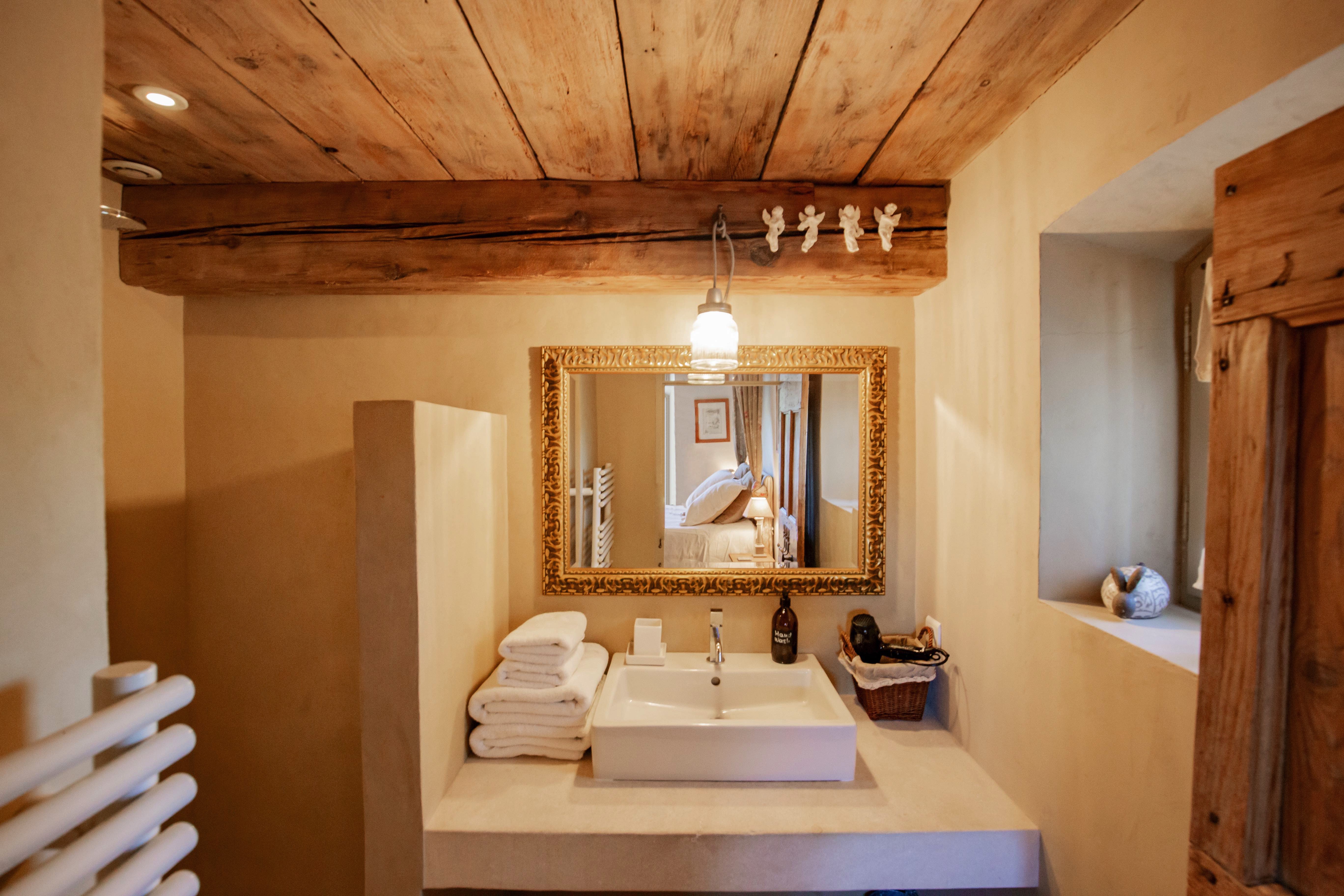 A cozy rustic bathroom featuring a white rectangular sink, ornate gold-framed mirror, wooden ceiling, beige walls, and hanging bulb lights. A basket and folded towels are displayed, adding to the warm ambiance.